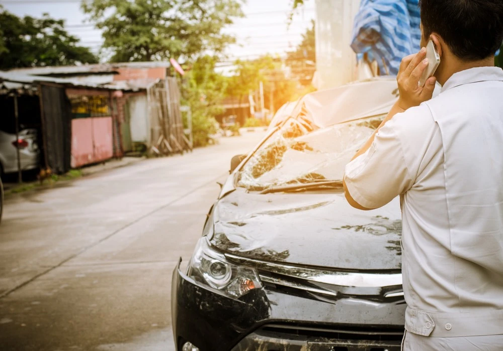 A person stands beside their damaged car, phone in hand, visibly concerned, highlighting the urgent need for vehicle repair following a car accident.